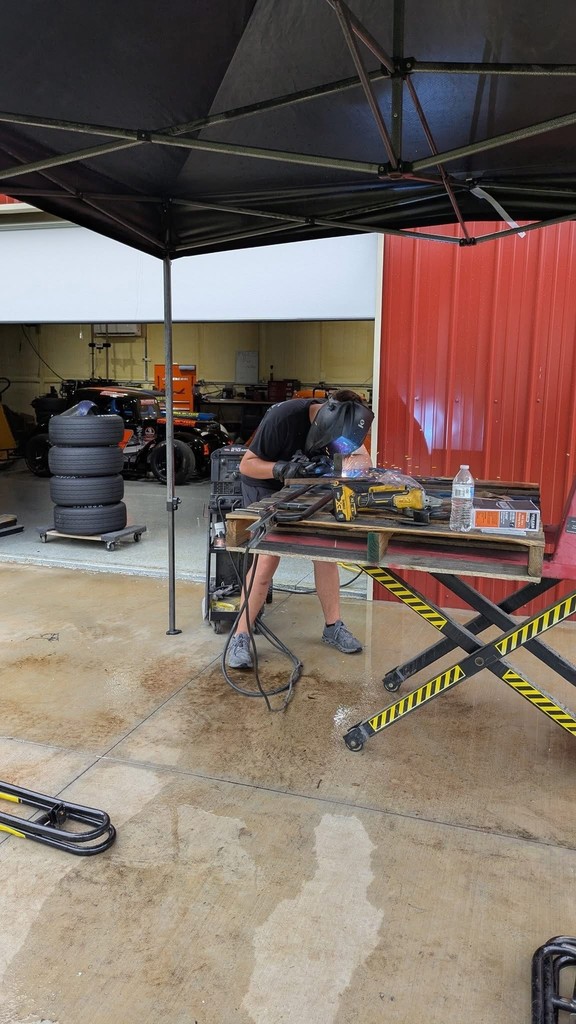 A student wearing protective gear and a welding helmet is welding metal parts on a workbench outside a garage. Several car tires are stacked nearby, and a race car is visible in the background. The scene shows hands-on mechanical work as part of an agricultural mechanics project.
