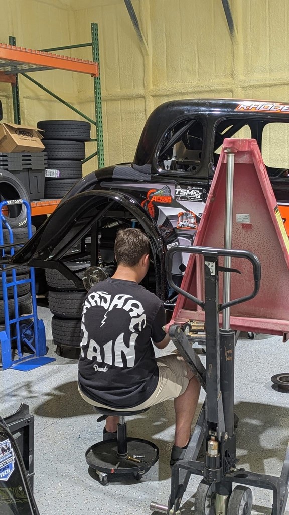 A student is seated in a garage working on a black Legends race car labeled “Rhodes.” The space has tools, car parts, and stacks of tires, showing the student performing maintenance and repair work related to his mechanical maintenance project.