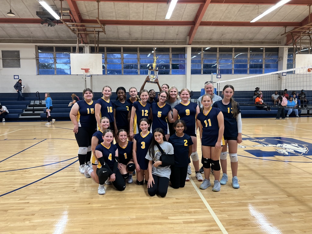 A middle school girls’ volleyball team poses together on a gym court after a game. The players, wearing navy and gold uniforms with numbers on the front, smile proudly as one player in the back holds up a large trophy. Two coaches stand behind the team, and the volleyball net and bleachers are visible in the background.