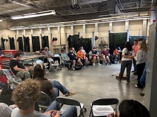 Students sit and listen to their peers presenting at the front of the agricultural shop during the FFA chapter meeting, with several students wearing orange and maroon sweatshirts