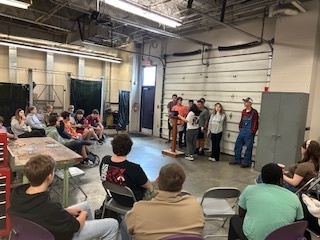 Students sit in chairs inside the agricultural shop while a group of FFA members stands at a podium giving announcements. Tools and welding booths line the room.