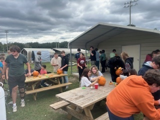 A large group of students gathers around picnic tables outside near the agriculture building, carving pumpkins and talking together during the FFA meeting.