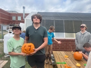Two students hold a carved pumpkin with the number seven on it while standing outside near a greenhouse. Other students work on their own pumpkins in the background.