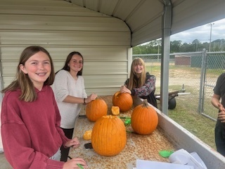 Three students stand at an outdoor table smiling as they carve pumpkins under a covered shelter. Several pumpkins are in progress, and tools are scattered on the table.