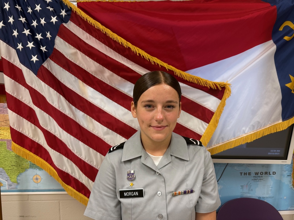 Hayden Morgan, a JROTC cadet wearing a gray uniform with name tag “Morgan,” stands in front of the American flag and the North Carolina state flag. She is smiling slightly, and a world map is visible on the wall behind her.