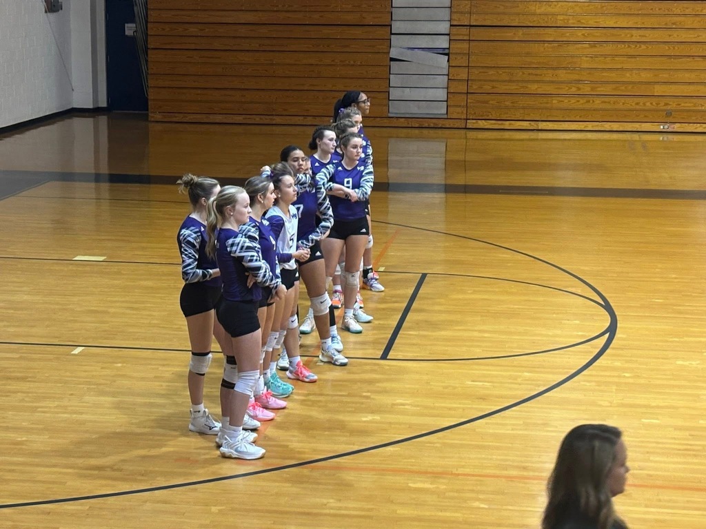 The Lady Knights volleyball team stands in a line on the court during a game, wearing matching purple and white uniforms. The players appear focused as they prepare for the match.