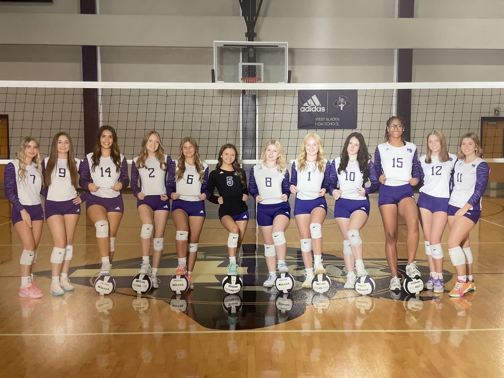 The Lady Knights volleyball team stands in a line on the court during a game, wearing matching purple and white uniforms. The players appear focused as they prepare for the match.