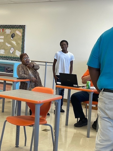 A student in a white shirt stands at a desk with a laptop, speaking to two adults seated nearby in a classroom with orange chairs.