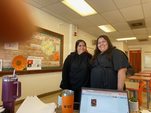 Two adults smile for the camera in a classroom with orange desks and colorful bulletin boards behind them.