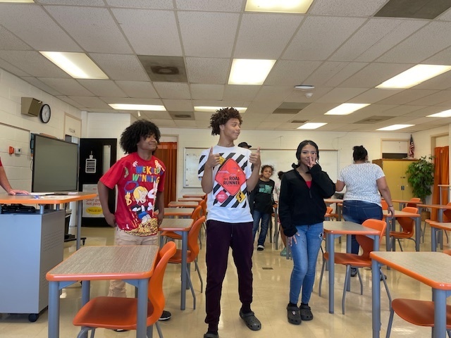 Three students stand at the front of a classroom smiling and talking, with orange desks and other students in the background.