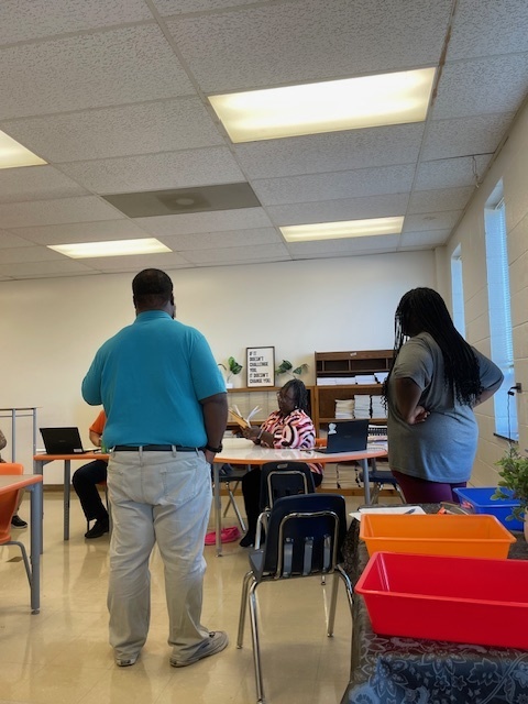 A student meets with family members and a teacher in a classroom. The adults are standing and sitting around tables, engaged in conversation.