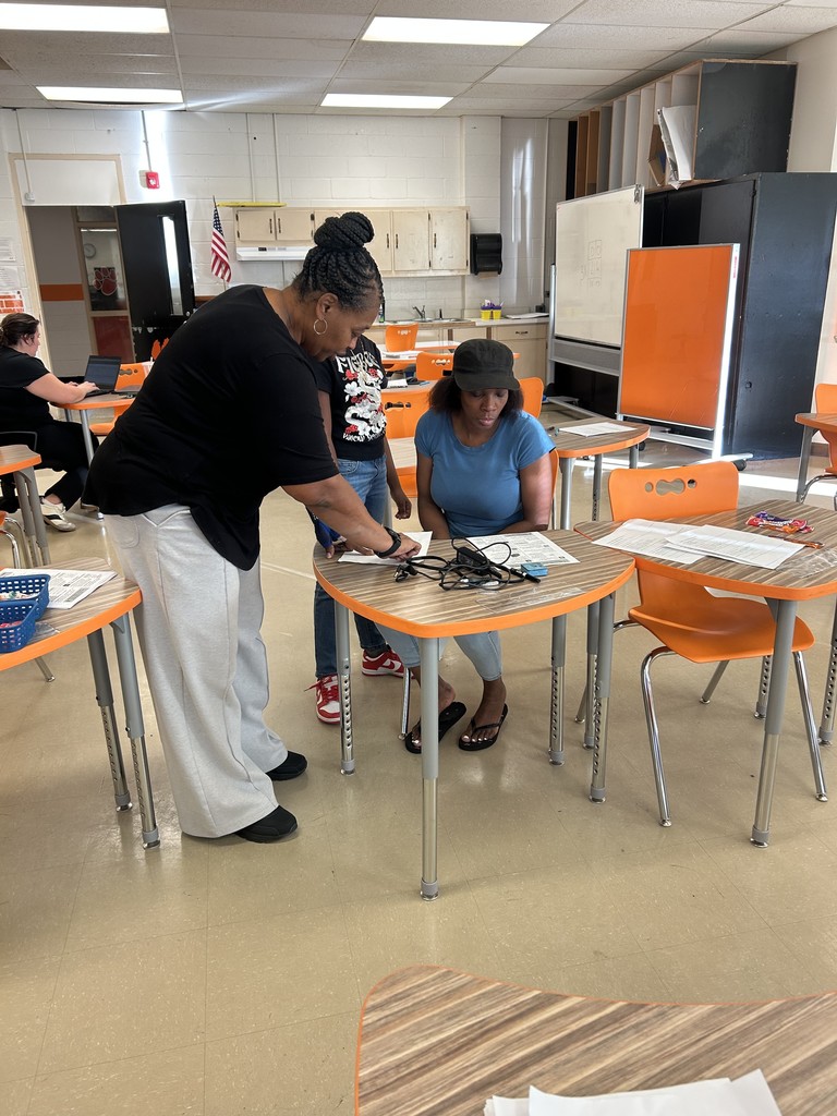 A teacher assists a student and family member seated at a classroom table during a student-led conference, with laptops and papers on the desk.