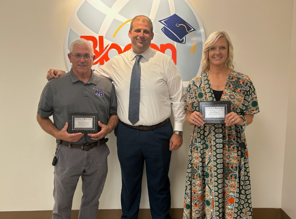 Coach Darryl Rutland, Superintendent Dr. Jason Atkinson, and Coach Nikky Bryant stand together in front of a Bladen County Schools logo. Coach Rutland and Coach Bryant each hold a framed certificate recognizing them for their heroism during a recent incident. All three are smiling for the photo.