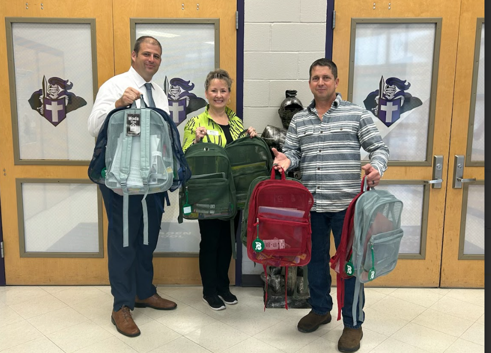Three adults stand in front of West Bladen High School doors holding donated backpacks filled with school supplies. The man on the left is wearing a white shirt and tie, the woman in the center is wearing a green and black patterned top, and the man on the right is wearing a striped shirt. The backpacks are red, green, and clear, and the West Bladen Knights logo is visible on the doors behind them.
