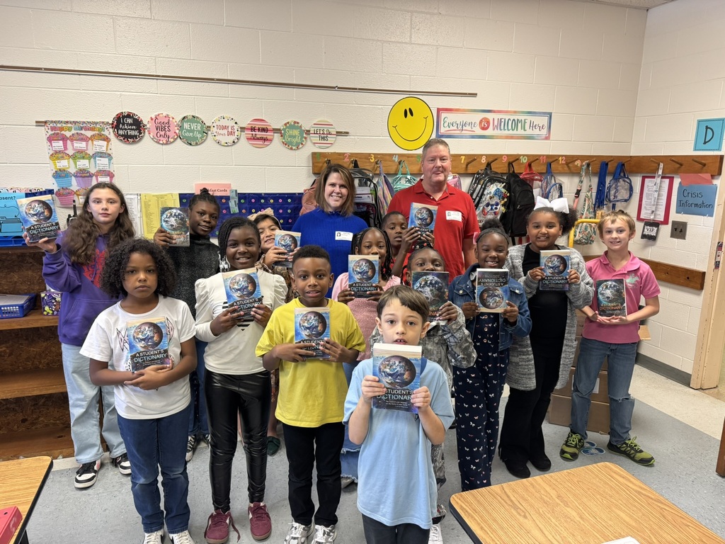 A group of third-grade students stand in their classroom holding new dictionaries with big smiles. Two Rotary Club members stand behind them, also smiling. The classroom wall is decorated with colorful motivational signs and student artwork, and backpacks hang along the back wall.