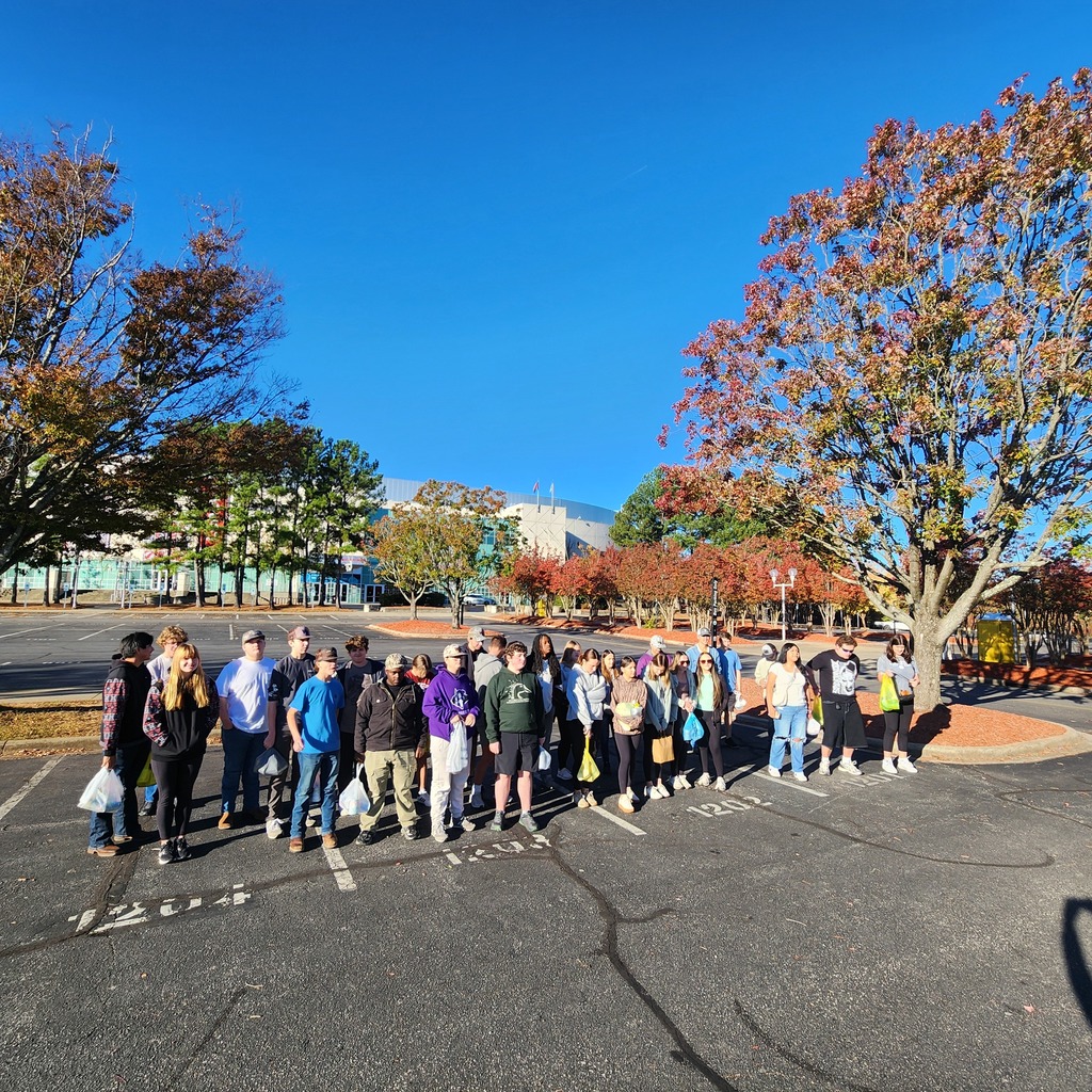 A group of high school students stands together in a parking lot on a sunny fall day, surrounded by colorful trees with red and orange leaves. They appear to be on a field trip or group outing, with a large modern building visible in the background under a clear blue sky.