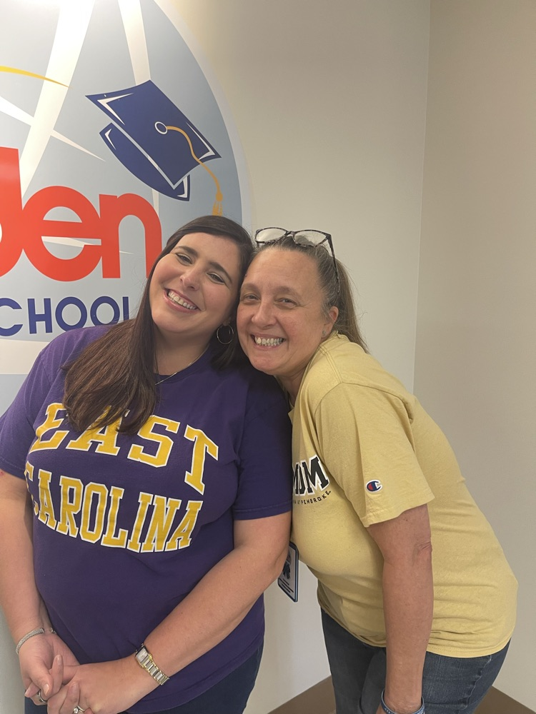 Two women smile while standing in front of a wall with the Bladen County Schools logo. One wears a purple “East Carolina” shirt and the other wears a yellow “UNCP Mom” shirt.