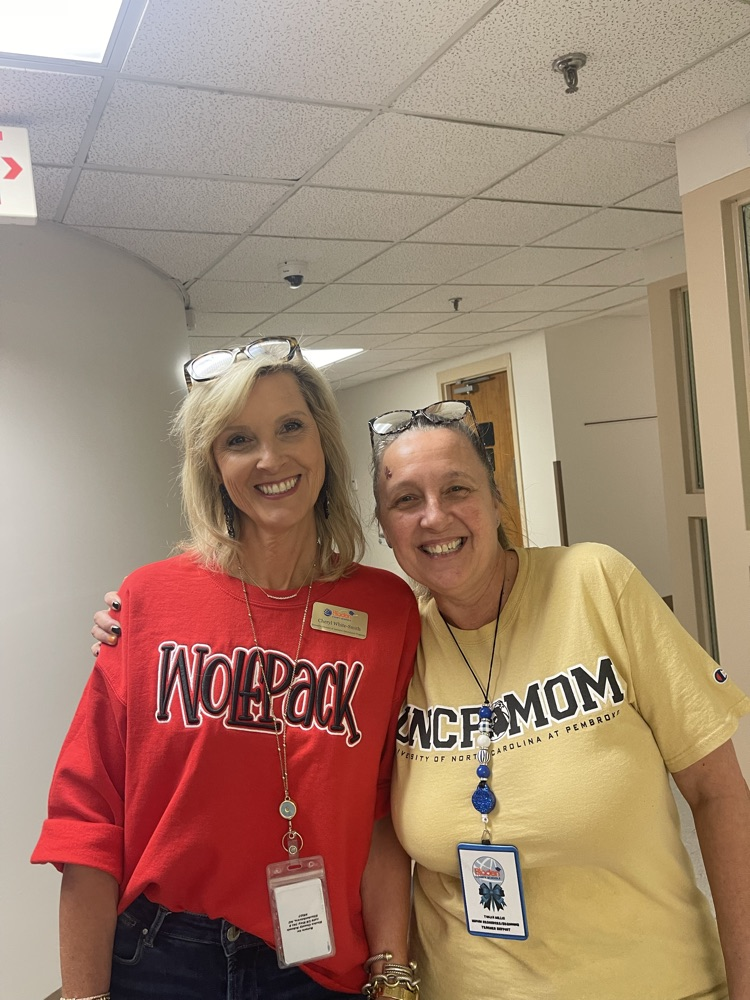 Two smiling women stand side by side in a hallway. One wears a red “Wolfpack” shirt and the other wears a yellow “UNCP Mom” shirt. Both have ID badges and glasses on their heads.