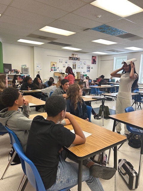 Students at Elizabethtown Middle School sit in small groups during a lively classroom activity. A teacher stands at the front of the room holding a paper as students look on and participate. In other shots, students are engaged in discussion while a teacher or staff member guides the activity. The classroom is bright and welcoming with colorful posters on the walls and papers spread across the desks as students collaborate and learn together.