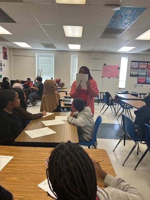 Students at Elizabethtown Middle School sit in small groups during a lively classroom activity. A teacher stands at the front of the room holding a paper as students look on and participate. In other shots, students are engaged in discussion while a teacher or staff member guides the activity. The classroom is bright and welcoming with colorful posters on the walls and papers spread across the desks as students collaborate and learn together.