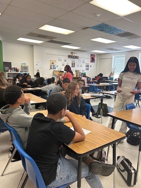 Students at Elizabethtown Middle School sit in small groups during a lively classroom activity. A teacher stands at the front of the room holding a paper as students look on and participate. In other shots, students are engaged in discussion while a teacher or staff member guides the activity. The classroom is bright and welcoming with colorful posters on the walls and papers spread across the desks as students collaborate and learn together.
