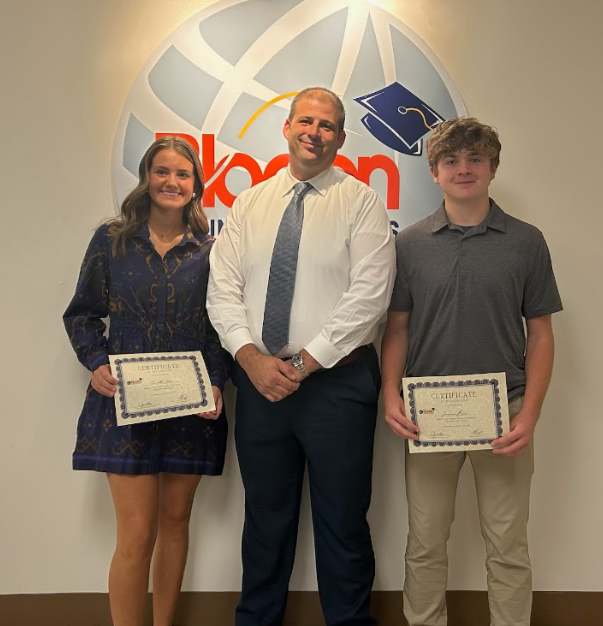 Two students stand with Superintendent Dr. Jason Atkinson in front of the Bladen County Schools logo. The students are holding framed certificates recognizing their achievements. The student on the left is wearing a navy patterned dress, and the student on the right is wearing a gray shirt and khaki pants. All three are smiling for the photo.