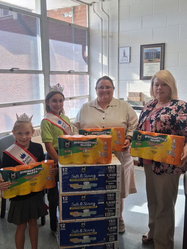 Two young girls wearing crowns and sashes, identified as Bladen County 4th of July Petite Miss Sadie Kelly and Junior Miss Rayne Kelly, stand with two adults inside Elizabethtown Primary School. They are smiling and holding large packs of disinfecting wipes while standing beside stacks of tissue boxes and cleaning supplies. Sunlight filters through large windows behind them, and framed pictures and motivational signs decorate the wall.