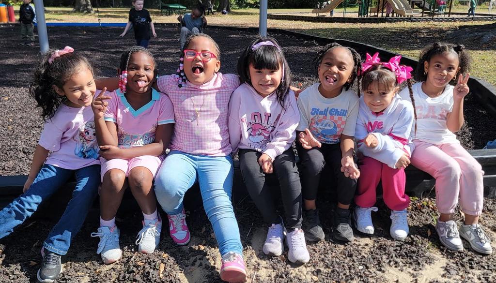 A group of seven young girls sit side by side on a playground border, smiling and making peace signs. They are dressed in shades of pink and white for Breast Cancer Awareness “Pink Out Day,” with playground equipment and classmates visible in the background.