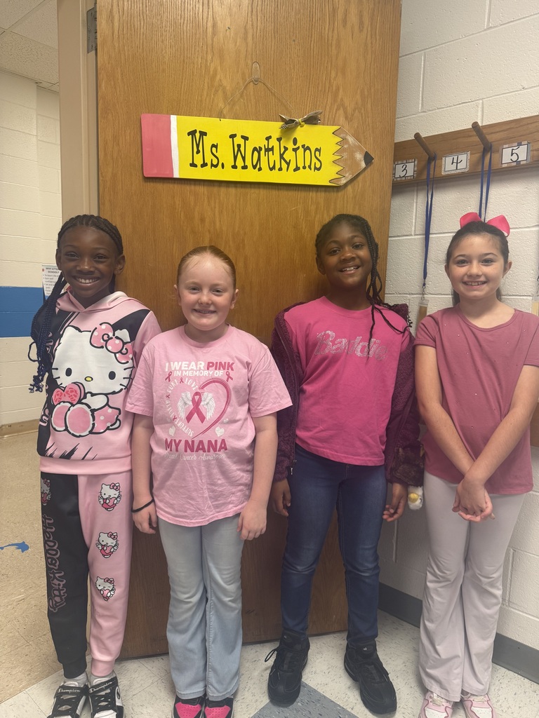 Four students stand in front of a classroom door with a “Ms. Watkins” sign above them. They are wearing pink shirts and outfits to show support for Breast Cancer Awareness “Pink Out Day.”