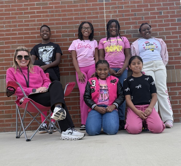 A teacher sits in a folding chair outside with six smiling students dressed in pink and black outfits for Breast Cancer Awareness Month. A brick wall serves as the backdrop.