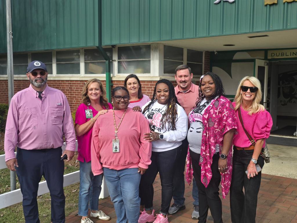A group of eight staff members stand together outside their school building, all wearing shades of pink in support of Breast Cancer Awareness Month.