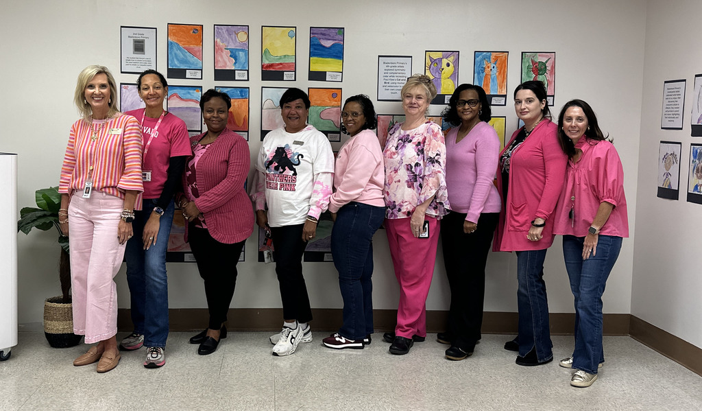 A group of nine staff members from Bladen County Schools stand together in a hallway decorated with colorful student artwork. Everyone is dressed in shades of pink for a districtwide Pink Out in recognition of Breast Cancer Awareness Month.