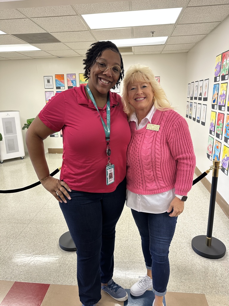 Two staff members stand side by side in a school hallway, both wearing pink sweaters and jeans to show support during Breast Cancer Awareness Month. Colorful student artwork is displayed on the wall behind them.