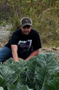 A person wearing a dark shirt and baseball cap crouches beside a large patch of leafy green plants in a garden or field, surrounded by tall wild vegetation in the background.
