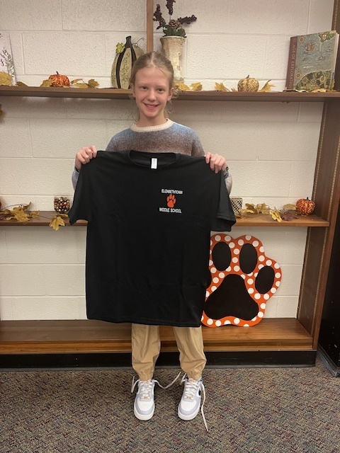 Olivia B., a smiling Elizabethtown Middle School student, stands indoors holding up a black ETMS spirit shirt