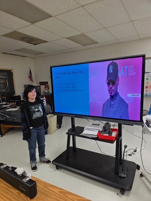 A student stands at the front of a classroom presenting a slideshow about Puerto Rican singer Farruko. The screen shows Farruko’s photo and biographical details.