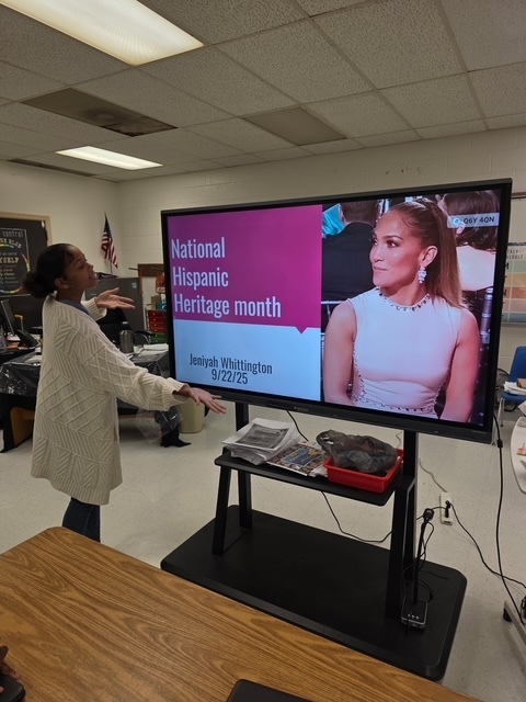 A student gestures toward a slide with Jennifer Lopez’s photo and the title “National Hispanic Heritage Month.” The screen includes the student’s name and date.