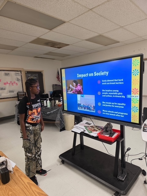 A student presents a slide titled “Impact on Society” featuring text and photos of Sonia Sotomayor. The student stands beside a bright blue slide that highlights her influence and accomplishments.