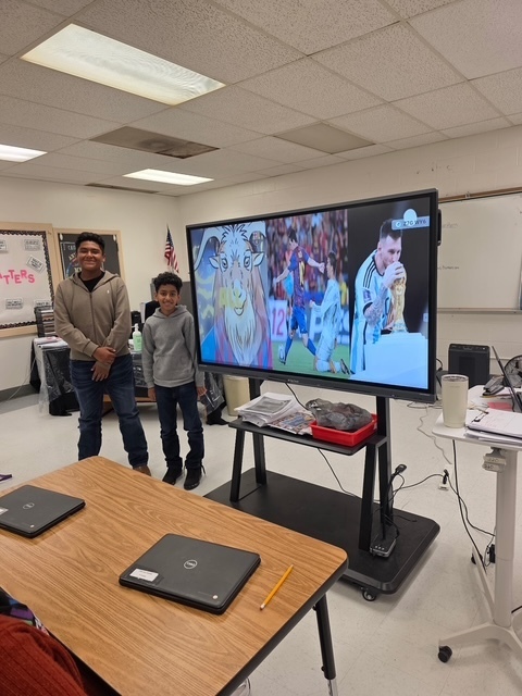 Two students stand beside a slide showing images of soccer star Lionel Messi and related artwork, smiling as they present.