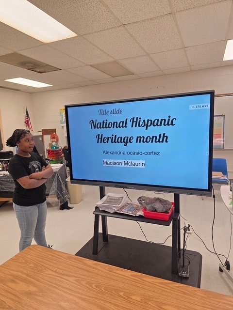 A student presents a slide titled “National Hispanic Heritage Month” featuring the names Alexandria Ocasio-Cortez and Madison McLaurin.