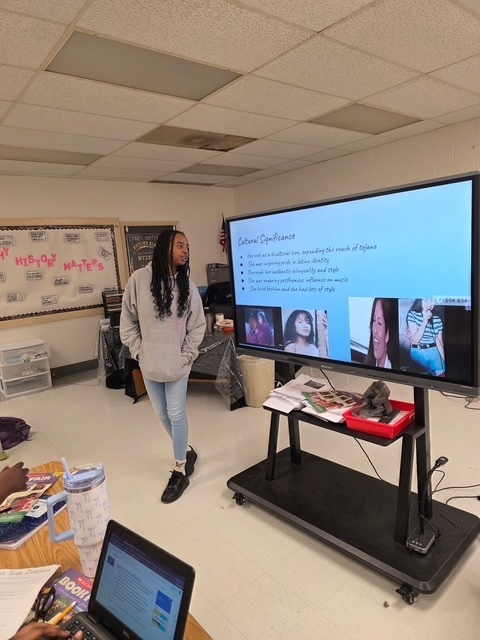 A student in a gray hoodie presents a slide about singer Selena. The screen shows multiple photos of Selena and text describing her cultural significance.