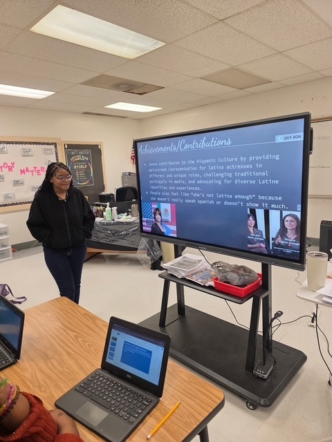 A student stands beside a slide discussing actress Jenna Ortega’s achievements and contributions to Hispanic culture. The presentation includes photos of Jenna Ortega.