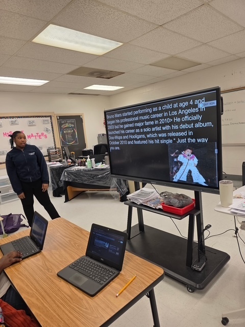 A student presents a slide about musician Bruno Mars, with a photo of him performing and text describing his early career.