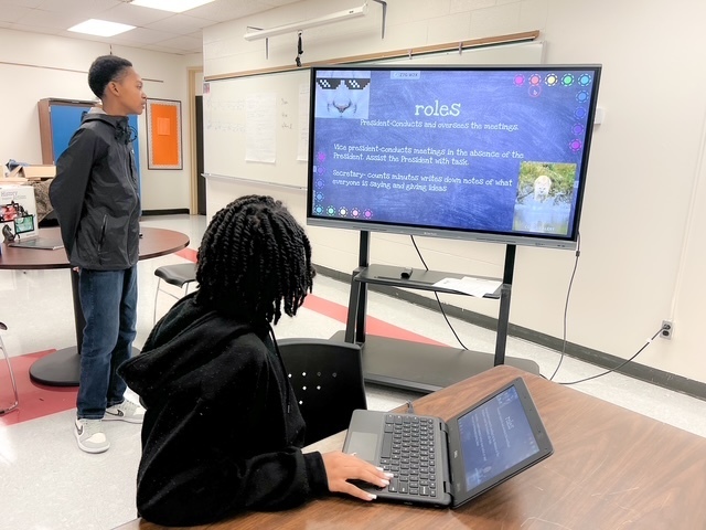 Two Elizabethtown Middle School students, Christopher H. and Jalaya L., present a slideshow on a large screen titled “Roles.” Jalyla sits at a laptop while Christopher stands nearby, both engaged in discussing their ideas for school improvement.