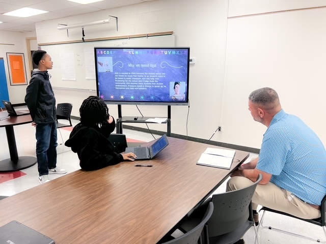 ETMS students Christopher H. and Jayla L. present their school improvement slideshow to Principal Mr. Godwin, who is seated at the table listening attentively. The presentation slide on the screen reads “Why We Need Elected Officials.