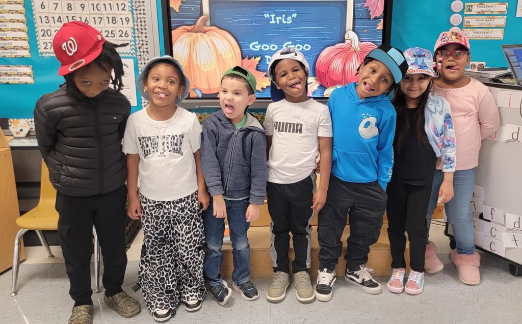 Eight young students stand in a classroom wearing a variety of fun hats, including baseball caps and bucket hats, while making silly faces and smiling. A display board behind them shows colorful pumpkins and the word “Iris.”