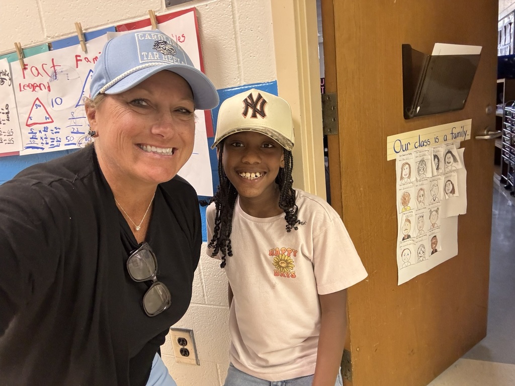 A teacher and a smiling student wearing baseball caps take a selfie together at the classroom door. A sign nearby reads “Our class is a family.”