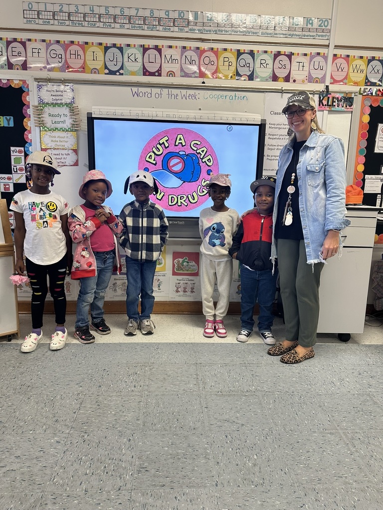 A teacher and six students stand in front of a classroom screen that reads “Put a Cap on Drugs.” Everyone is smiling and wearing different styles of hats to celebrate Red Ribbon Week.