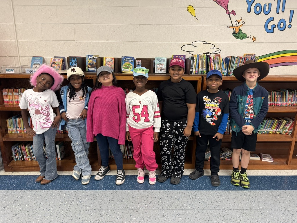 Seven students pose together in the library wearing hats of all kinds—cowboy, baseball, fuzzy pink, and more—while standing in front of a colorful bookshelf full of books.