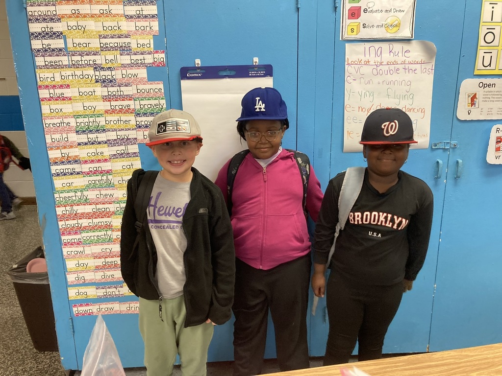 Three students pose together in front of a classroom word wall filled with vocabulary cards. Each wears a baseball cap, smiling and showing off their school pride.